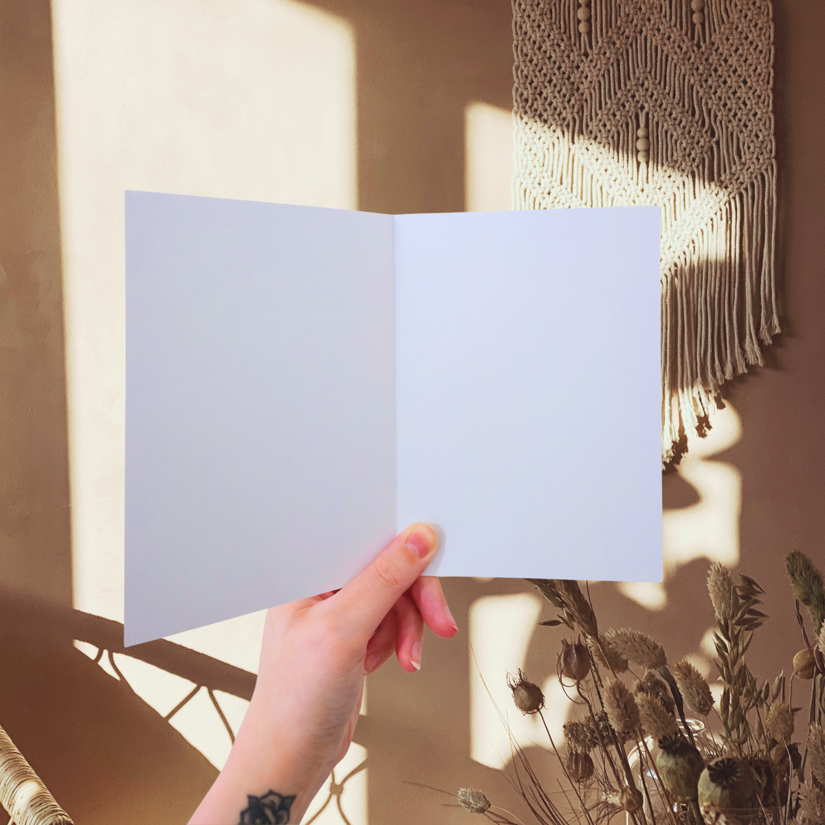 Hand holding a blank white greeting card with macrame art and plants in the background