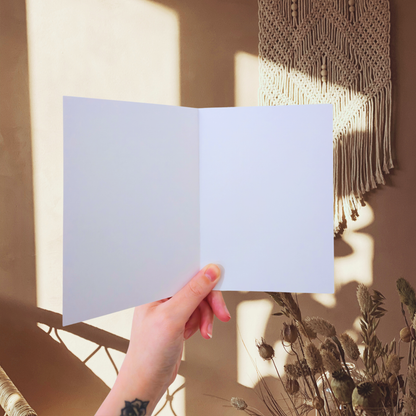Hand holding a blank white greeting card with macrame art and plants in the background