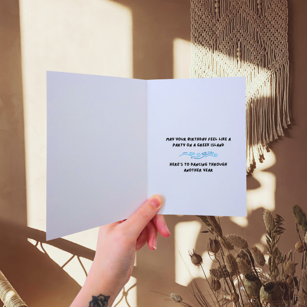 Hand holding a birthday card with text against a warm-toned wall with macrame art.