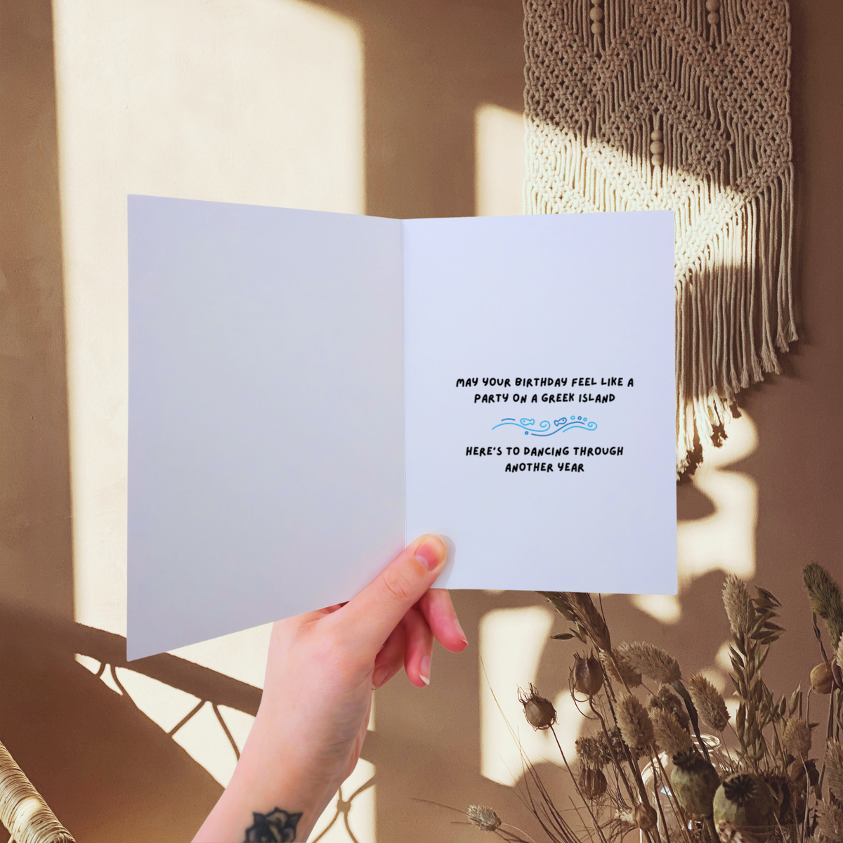 Hand holding a birthday card with text against a warm-toned wall with macrame art.