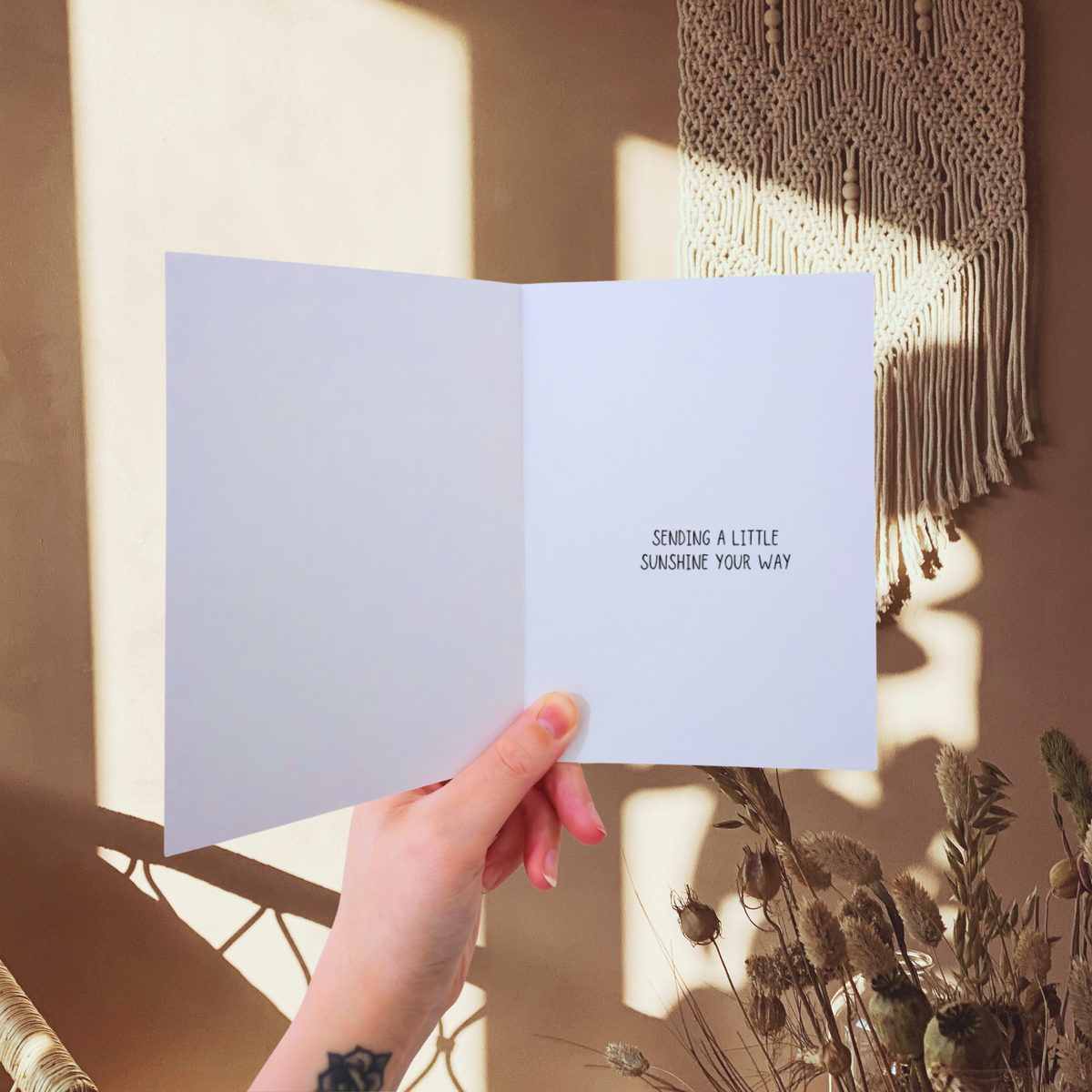 Hand holding a card with 'Sending a little sunshine your way' text, sunlit room with macrame wall hanging.