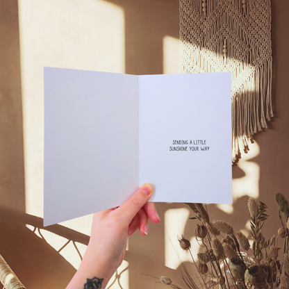 Hand holding a card with 'Sending a little sunshine your way' text, sunlit room with macrame wall hanging.
