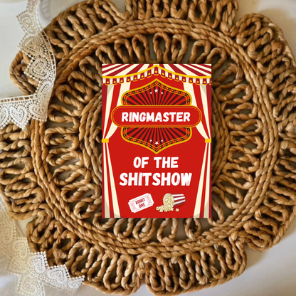 Red and white card with 'Ringmaster of the Shitshow' text on a woven mat.