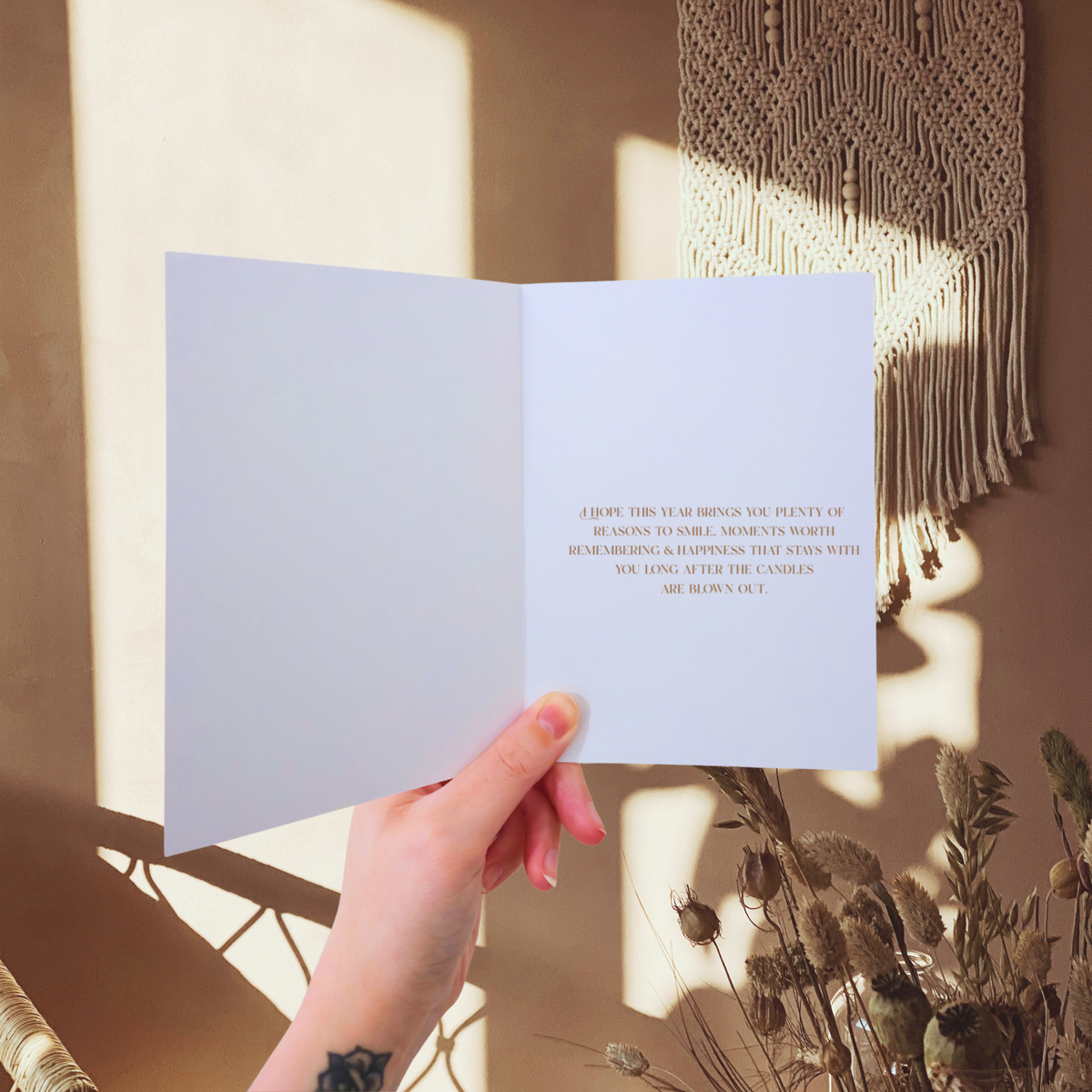 Hand holding a greeting card with a cat illustration and text, with a blurred background of plants and wall art.