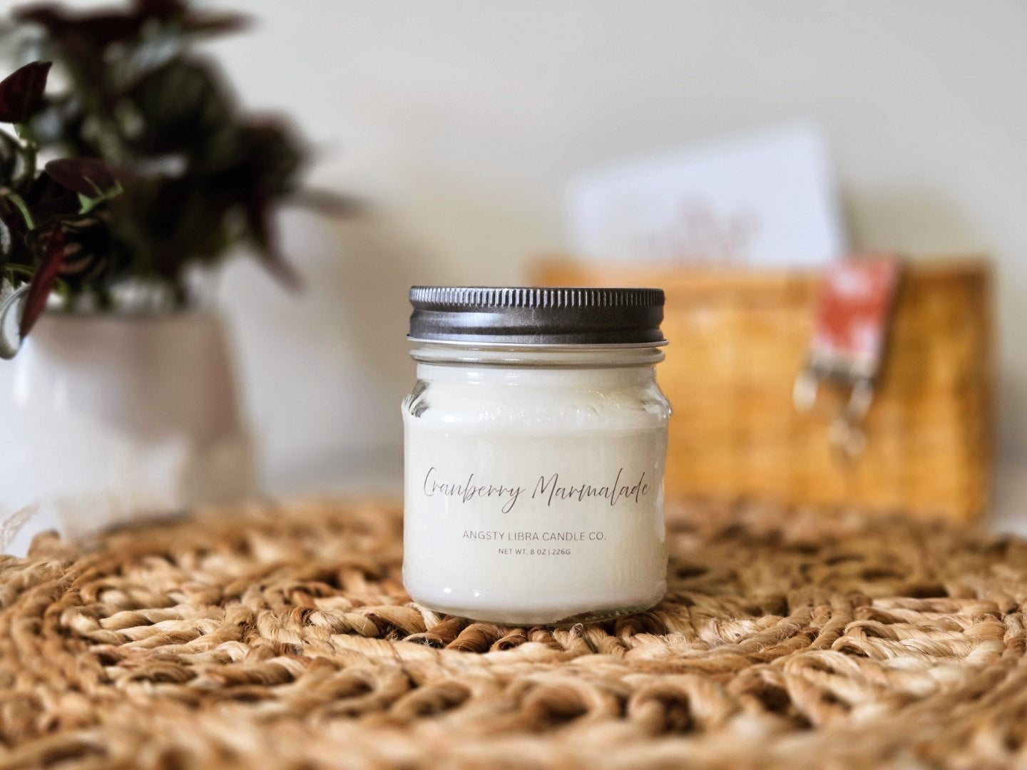Cranberry Apple Marmalade candle in a clear mason jar sitting on a wicker placemat with a blurred basket and greeting card in the background.